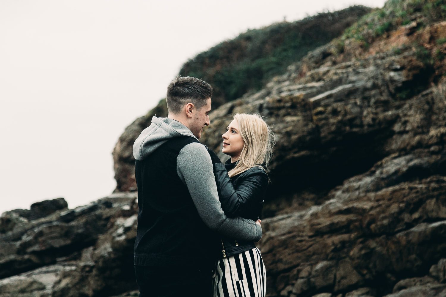 Bide and groom look into each others eyes standing near cliff on seaside engagement shoot